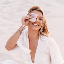 Load image into Gallery viewer, Happy blonde smiling model at the beach holding linen pouch over her eye wearing crystal 925 bracelets
