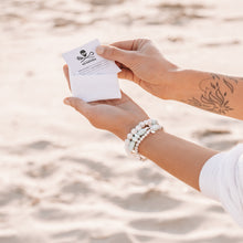 Load image into Gallery viewer, Two hands holding a white bracelet pouch with three howlite 925 sterling silver and peruvian opal bracelets on wrist and sand in the background
